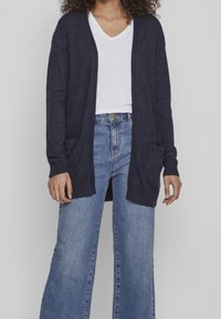 Cárdigan navy de tejido suave, diseño de abertura frontal con puños acanalados, que cuenta con dos bolsillos delanteros y se combina con unos vaqueros de pierna ancha en azul claro.