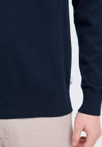 Suéter de punto azul marino con puños y dobladillo acanalados. Textura suave con una pequeña etiqueta de marca en el lateral. Pantalones beige visibles en la parte inferior.