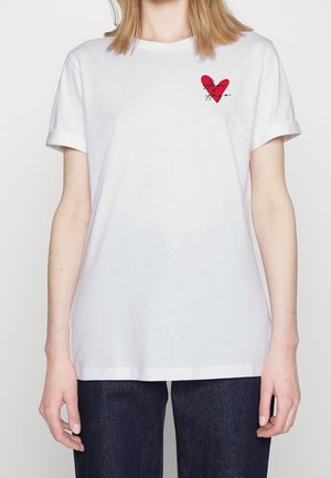 Osoba nosząca białą koszulkę z krótkim rękawem z małym czerwonym sercem i czarnym napisem na lewej piersi, połączoną z ciemnoniebieskimi dżinsami.