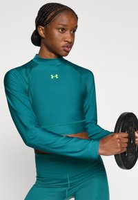 Μπλε πράσινο crop top με μακριά μανίκια και ψεύτικο λαιμό, σετ με αντίστοιχο ψηλόμεσο κολάν. Ξεχωρίζει το λογότυπο Under Armour σε φθορίζον κίτρινο.