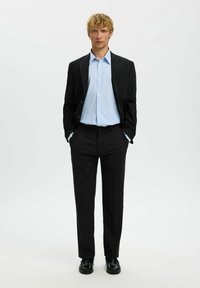 Zwarte colbert over een lichtblauw gestreept shirt, gecombineerd met zwarte broeken en schoenen, gepresenteerd tegen een witte achtergrond.