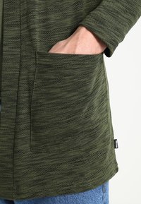 Maglietta a maniche lunghe verde oliva con un motivo testurizzato. Presenta una tasca laterale. Realizzata in tessuto morbido e leggero. L'orlo è leggermente curvato.