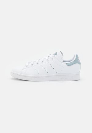 Biały sneaker Adidas Stan Smith z jasnoniebieską patką na pięcie i perforowanymi bocznymi paskami, pokazany w widoku bocznym na jednolitym tle.
