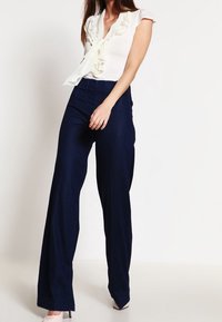Bílé tričko s volánovými detaily a šňůrkou u límce, kombinované s vysokým pasem a širokými modrými kalhotami. Hladká textura, přiléhavý pas.