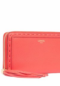 Portefeuille en cuir corail avec une fermeture zippée, mettant en valeur des coutures décoratives et un accent en pompon. Logo doré "Lancel Paris" embossé à l'avant.