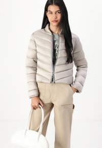 Light beige puffer jacket with horizontal quilted design, zip closure, worn over a graphic t-shirt and paired with tan trousers and a white handbag.