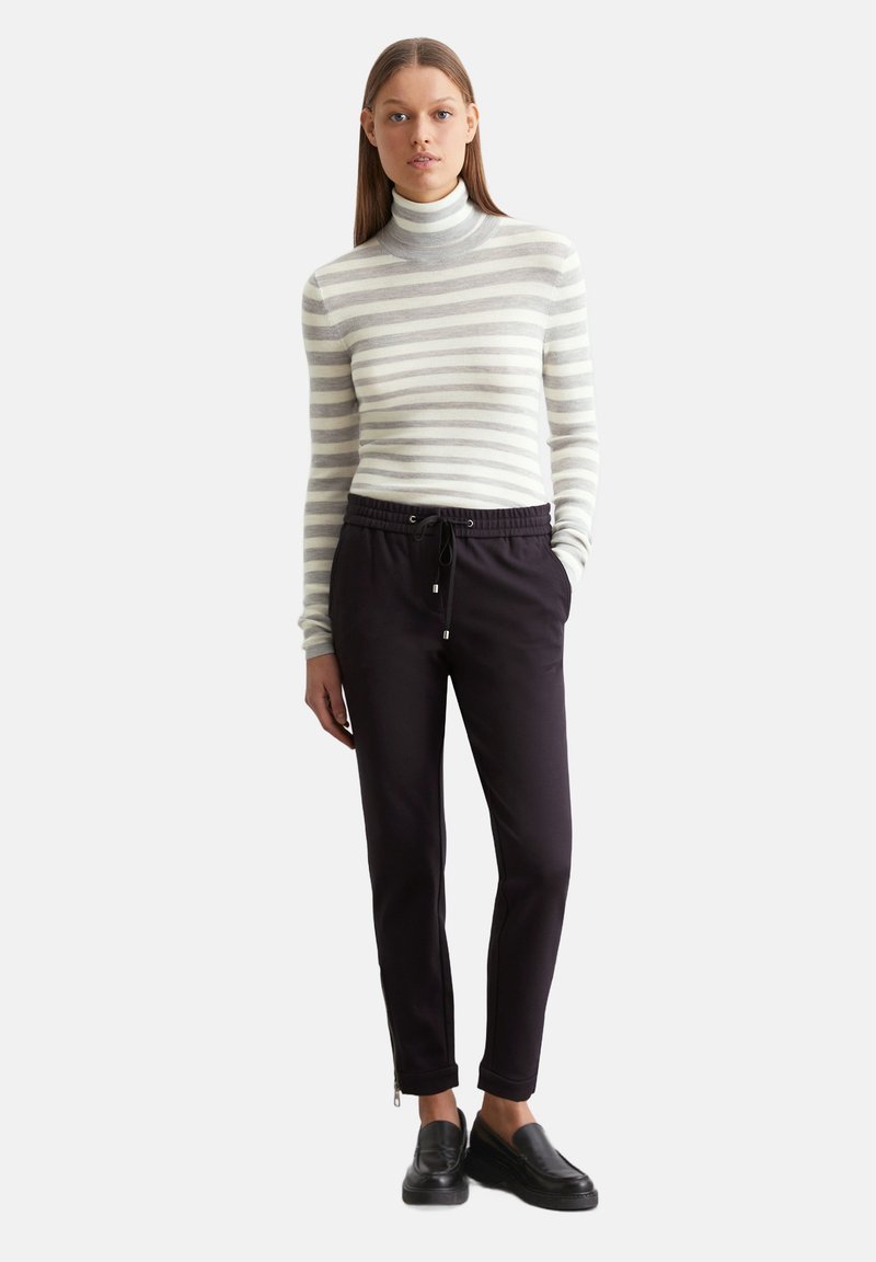 Gestreifter Rollkragenpullover in Hellgrau und Weiß, kombiniert mit dunklen Hosen mit elastischem Bund und Reißverschluss an den Knöcheln. Schwarze Slipper.