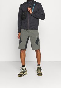 Schwarze Herren-Zip-Jacke mit blauen Akzenten, graue Shorts mit dunklen geometrischen Mustern und Wanderschuhe mit neongelben Details.