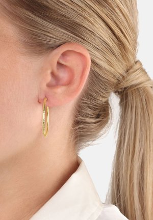 Gouden hoopoorbel met een geslagen textuur, cirkelvorm, en een gepolijste afwerking, gedragen in het linkeroor van een persoon met blond haar.