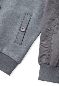 Sudadera gris con un bolsillo abotonado, que presenta una tela texturizada. A su lado hay una chaqueta gris oscuro con un aspecto acolchado y arrugado.
