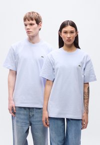 Μαλακά βαμβακερά t-shirt σε ανοιχτό μωβ χρώμα με κοντά μανίκια, με μικρό πράσινο κεντημένο λογότυπο στο στήθος, συνδυασμένα με ξεθωριασμένο μπλε τζιν.