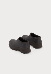 Pantofi de cauciuc maro cu o suprafață netedă și texturată, formă rotundă și o talpă groasă. Au un design slip-on cu o guleră joasă.
