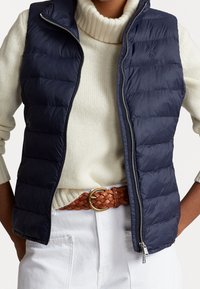 Modellen bär en marinblå pufferväst över en krämvit tröja, matchad med ett flätat brunt bälte och vita byxor, vilket visar upp en avslappnad höstoutfit.