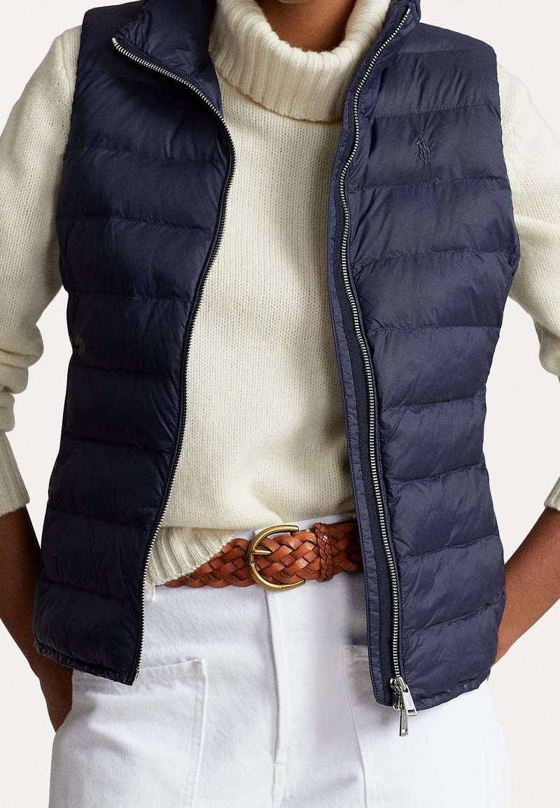 Modellen bär en marinblå pufferväst över en krämvit tröja, matchad med ett flätat brunt bälte och vita byxor, vilket visar upp en avslappnad höstoutfit.