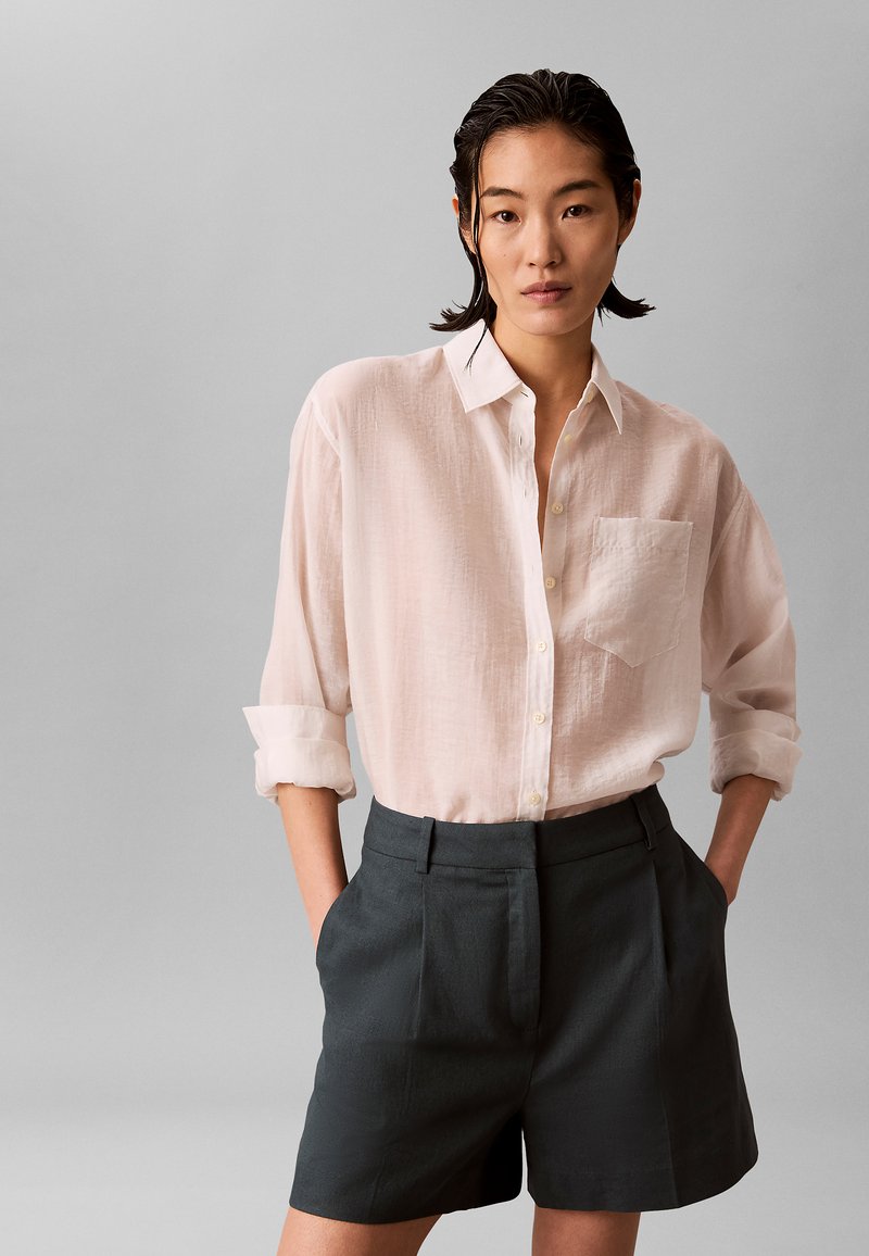 Femme aux cheveux courts et foncés porte une chemise rose clair boutonnée avec des manches retroussées et un short foncé taille haute, debout devant un fond gris.