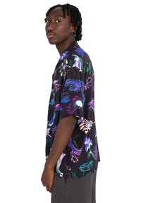 Camisa negra de manga corta con estampados florales coloridos en tonos de azul, rosa, verde y blanco. Diseño informal con un ajuste relajado.