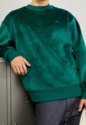 Person, die einen dunkelgrünen samtigen Pullover trägt und sich an graue Spinde lehnt, hält ein helles Tuch in einer Hand, blaue Jeans sichtbar.
