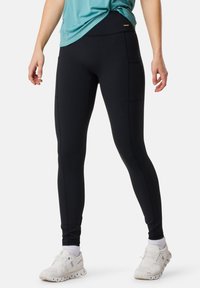 Leggings noirs taille haute, en tissu extensible, avec des poches latérales et des détails de couture contrastants, assortis à des chaussures de sport blanches.