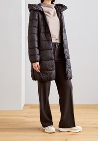 Černý prošívaný péřový kabát s kapucí, s prošitým vzorem, uzavíratelný na zip a s kapsami. Kombinovaný s béžovým topem a tmavými kalhotami.