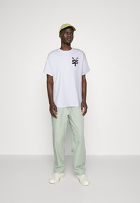 Hvid bomulds-T-shirt med et sort grafisk design på brystet, kombineret med lysegrønne bukser og en struktureret grøn cap. Hvide sneakers.