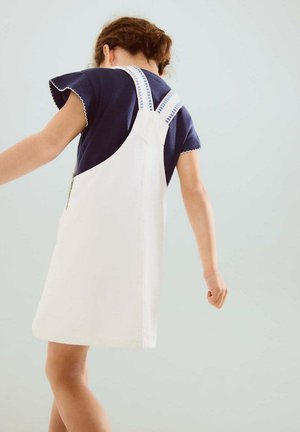 Copil cu păr castaniu purtând o cămașă bleumarin și o rochie albă tip șorț cu bretele albastre în dungi, stând într-un fundal simplu și deschis la culoare.