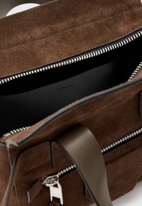 Borsa in suede marrone con chiusura a zip, hardware in argento e fodera interna nera. Presenta una tracolla piatta e una superficie testurizzata.