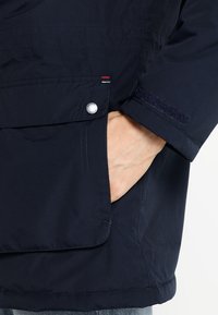 Main placée dans la poche d'une veste bleu marine avec rabat, bouton argenté et bracelet en Velcro au niveau du poignet.