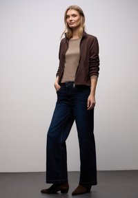 Brązowa bomberka zamszowa na beżowej bluzce, w połączeniu z ciemnymi dżinsami o szerokich nogawkach i brązowymi botkami za kostkę. Luźny krój i casualowy design.