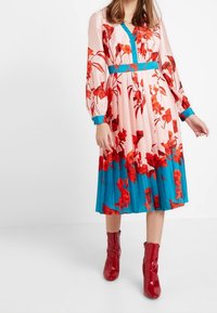 Blumen-Midi-Kleid in Rosa und Türkis mit langen Ärmeln, V-Ausschnitt und taillierter Taille. Mit lebhaften roten Blumenmustern und Falten.