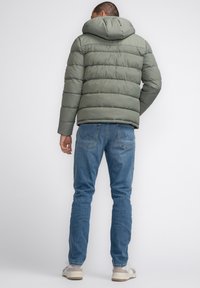 Chaqueta de plumas verde con costuras horizontales, capucha adjunta y puños elásticos. Llevada sobre unos vaqueros azules y zapatillas de color claro.