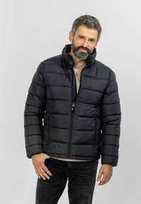 Black puffer jacket with a zip closure, featuring a high collar and horizontal stitched detailing. Worn over a beige shirt and black pants.