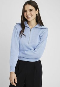 Pull en tricot bleu clair avec un col zippé et des manches longues. Poignets et ourlet côtelés, porté avec un pantalon noir. Texture lisse et design ajusté.