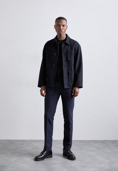 Navy wool-blend jacket with four front pockets, button closure, and long sleeves paired with dark trousers and black shoes on a textured floor.