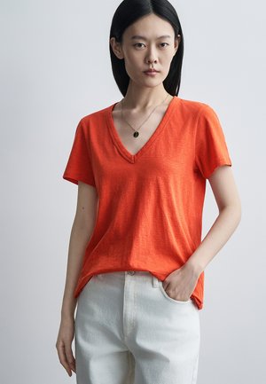 Kobieta z prostymi czarnymi włosami ubrana w pomarańczową koszulkę z krótkim rękawem i dekoltem w serek oraz białe spodnie, stoi z jedną ręką w kieszeni na jednolitym tle.