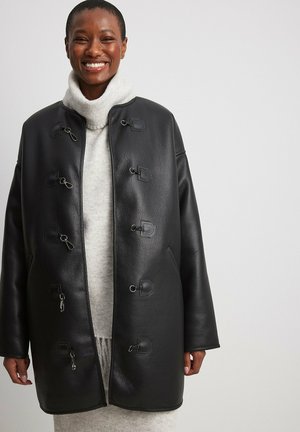 Cappotto in pelle nera con una vestibilità rilassata, chiusure a cordoncino e colletto alto. Indossato sopra un maglione grigio chiaro, con una texture liscia.