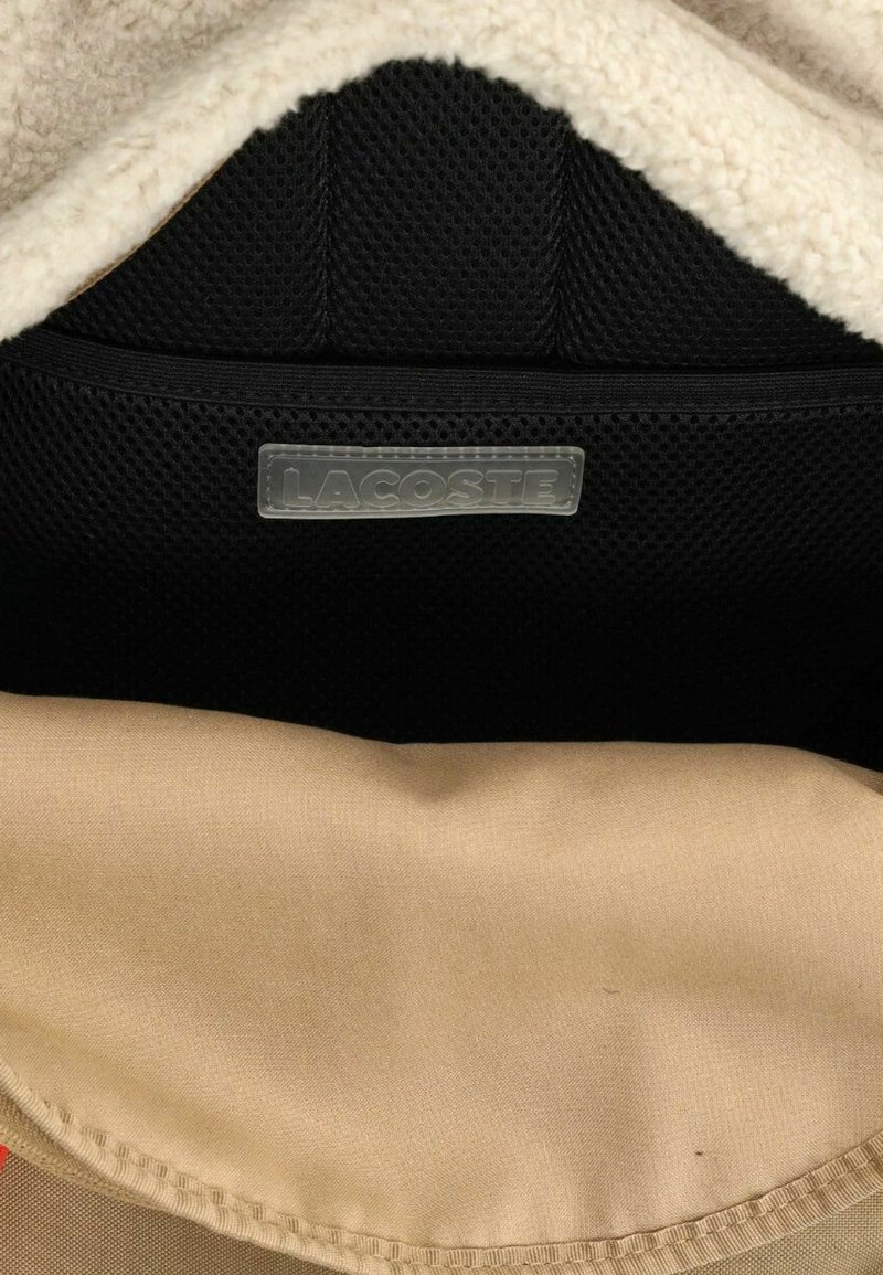Interior de una bolsa con un suave forro de franela crema, un panel de malla negra y una base de tela marrón con una etiqueta del logo de Lacoste clara.