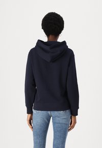 Haut à capuche bleu marine avec une capuche spacieuse, des poignets et un ourlet côtelés ; en mélange de coton doux. Porté avec un jean bleu clair. Design minimal.