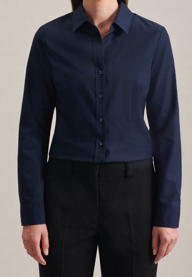 Femme portant une chemise cintrée bleu marine à boutons avec manches longues et un pantalon noir sur mesure, debout devant un fond uni.