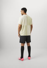T-shirt à manches courtes de couleur verte claire avec un design coloré et un petit logo dans le dos, associé à des shorts noirs et des crampons de football roses.