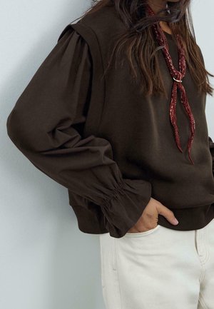 Femme portant un haut à manches bouffantes marron foncé avec un foulard rouge autour du cou et un pantalon clair, la main dans la poche, adossée à un mur clair.