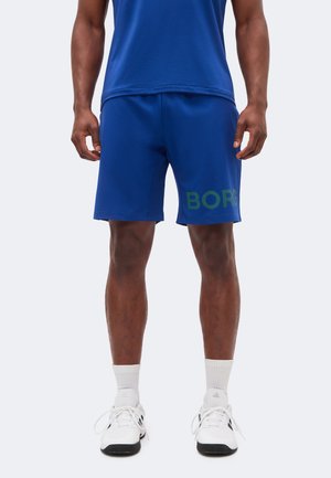 Muškarac u plavim kratkim hlačama s tekstom "BORG" na lijevoj nozi, bijelim čarapama i bijelim sportskim cipelama stoji nasuprot jednobojnoj pozadini.