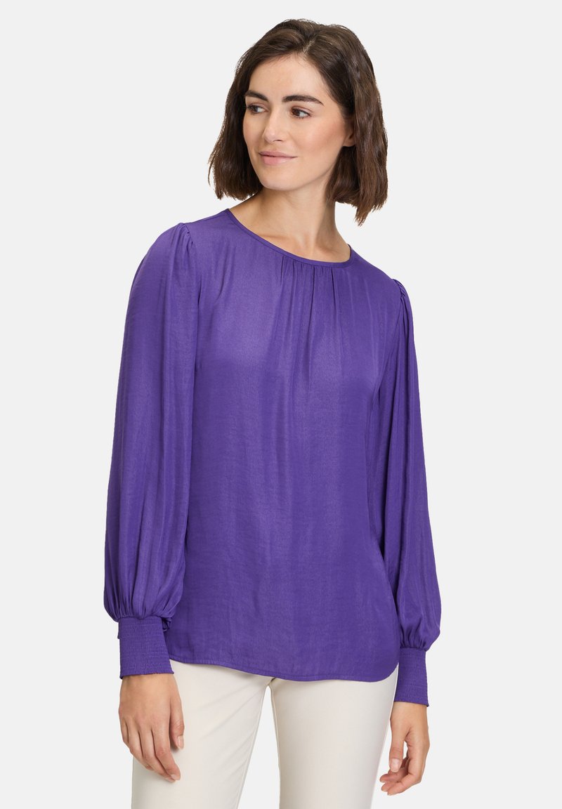 Lilla langærmet bluse med rund halsudskæring, plisseret front og samlet ærmer. Stoffet ser let og en smule gennemsigtigt ud.