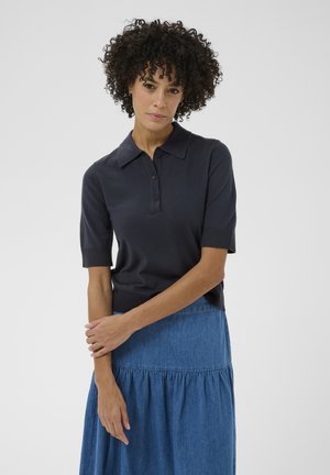 Kobieta o kręconych włosach, ubrana w ciemną, krótką koszulkę polo i niebieską dżinsową spódnicę, stojąca z założonymi rękami na tle jednolitej ściany.