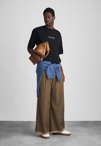 Zwarte t-shirt met witte tekst, blauwe sweater om de taille geknoopt, bruine wijde broek, bruine tas en witte sneakers, op een grijze achtergrond.