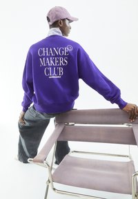 Paarse sweatshirt met witte tekst "CHANGE MAKERS CLUB" op de achterkant, gecombineerd met donkergrijze baggy jeans en een lichtpaarse cap.