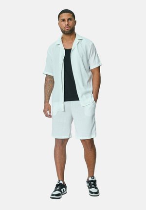 Chemise blanche à manches courtes et short assorti avec un motif texturé, accompagnés d'un débardeur noir et de baskets noires.