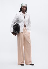 Γυναίκα που φοράει λευκό κοντό πουκάμισο δεμένο, μπεζ παντελόνια φαρδιάς γραμμής, μαύρα παπούτσια, παστέλ μαντίλι με σχέδια στο κεφάλι, κρατώντας μια μεγάλη μαύρη τσάντα.