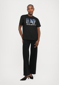 Persona, kas ģērbjas melnās plašās biksēs, melnos norādītājus papēžos un melnā T-kreklā ar zilu un sudraba EA7 Emporio Armani logo.