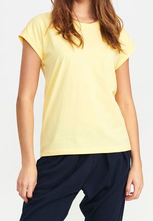 Kvinde iført en kortærmet gul T-shirt og løstsiddende mørkeblå bukser, stående foran en ensfarvet lys baggrund.