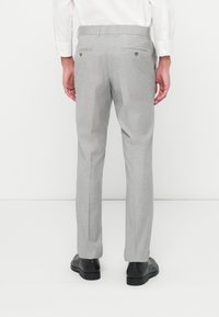 Calças cinza claro, ajustadas, com um acabamento texturizado. Apresenta dois bolsos traseiros com fechos de botão, usadas com sapatos pretos.